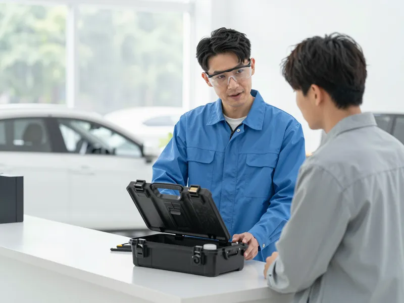 Ein Kfz-Glastechniker erklärt einem Kunden einen Prozess an einem Werkzeugkoffer neben einem Auto.